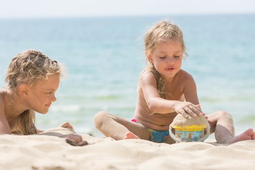 Two girls on the beach on a sunny day, playing with sand and a bucket on a background of the sea