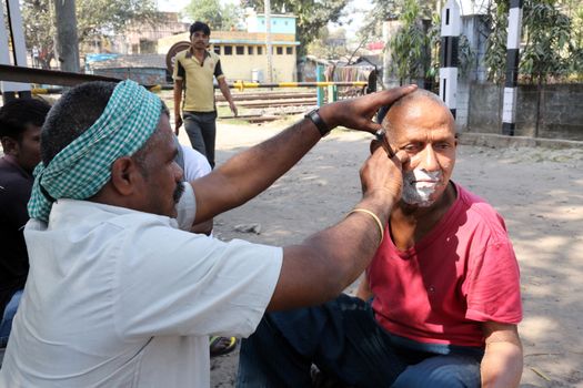 Street barber shaving a man using an open razor blade on a street in Kolkata, West Bengal, India