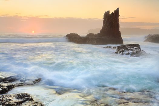 The new day sun glimpses over the horizon at Cathedral Rock, Kiama, Australia.  A sea mist or fog obscures clarity but softens the scene.