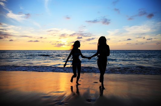 Two young happy women walking on tropical sea beach at sunset