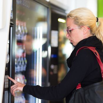 Casual caucasian woman using a modern beverage vending machine. Her hand is placed on the dial pad and she is looking on the small display screen.