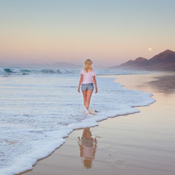 Woman walking on sandy beach in sunset. Waves sweeping away her traces in sand. Beach, travel, concept. Copy space. Square composition.