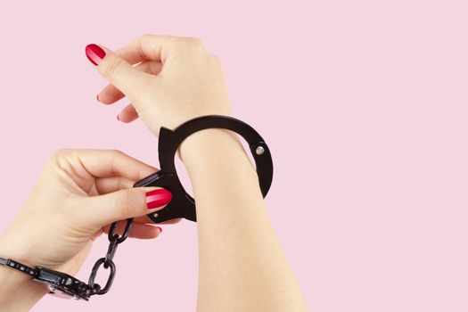 Beautiful female hands in black handcuffs isolated on pink background. Submission, trust and passion concept. 