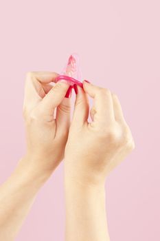 Female hands with red nails holding a pink condom isolated on pink background. Safe sex and birth control concept. 