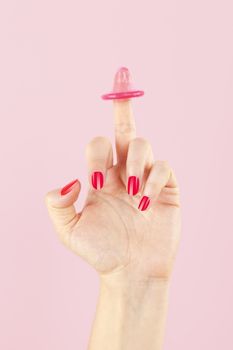 Female hands with a pink condom on middle finger isolated on pink background. Safe sex and birth control concept. 