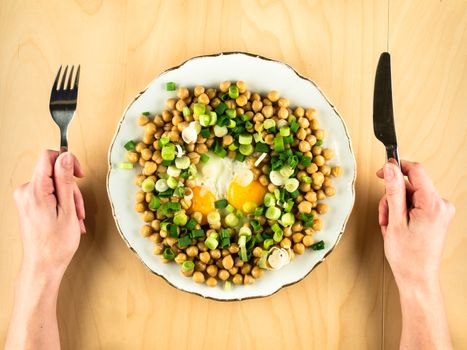 Plate with chick peas, fried eggs, chopped fresh onion with hands holding fork and knife, healthy eating, simple top view, copy space