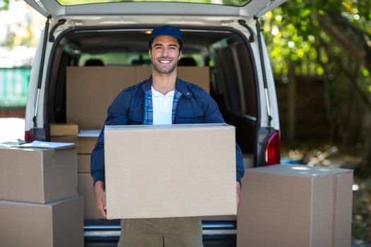 Portrait of smiling delivery man carrying cardboard box while walking by van