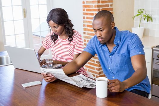 Young woman using laptop in the kitchen and man reading newspaper