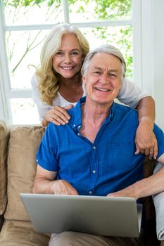 Portrait of senior couple with laptop sitting on sofa against window at home