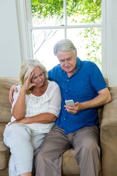 Serious senior couple using mobile phone while sitting on sofa at home