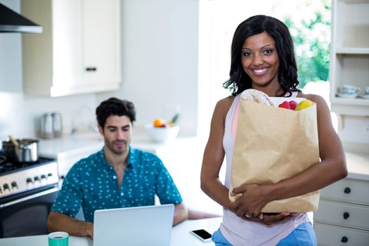 Portrait of woman with a bag of groceries and man working on laptop in background