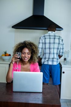 Woman using laptop and having wine while man cooking food in background at kitchen