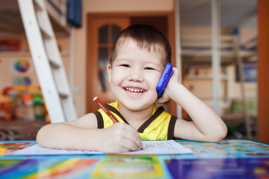 Happy little boy talking on smartphone at home while writing letters, three years old.