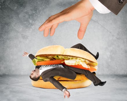 Businessman inside hamburger and big humand hand on grey wall background