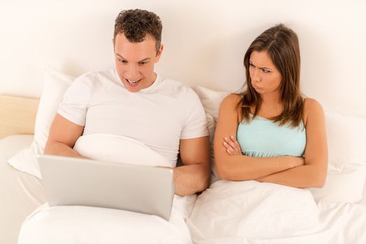 Couple with laptop in bed. Happiness man surfing on the net at laptop, woman upset and angry looking at him. 