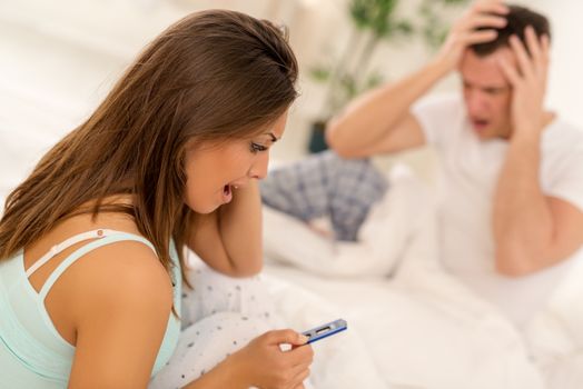 Young woman sitting on the bed and worried looking at pregnancy test. In the background is her desperate partner.