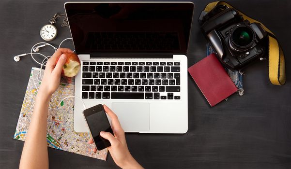 Outfit of the traveler. The view from the top. young woman sitting at the vintage brown wooden table. there are the map, camera, passport, watch, laptop and mobile phone with headphones on the table 