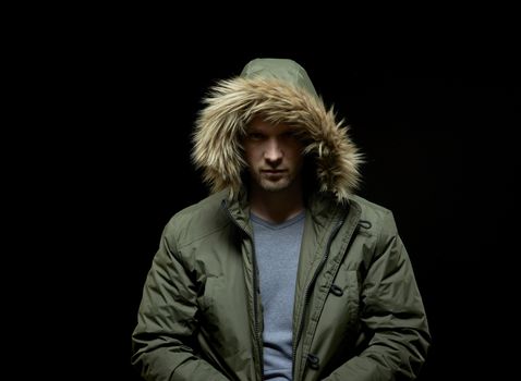 Low key studio portrait of suspicious young adult caucasian model wearing winter coat with hood on. Isolated on black.
