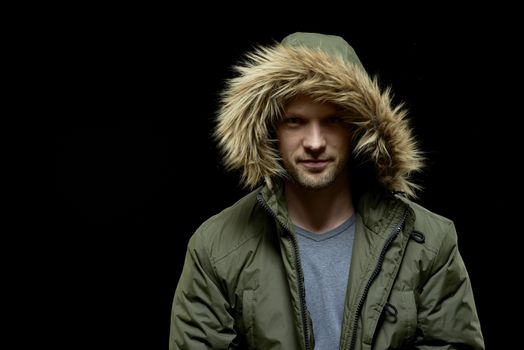 Low key studio portrait of young adult confident caucasian model wearing winter coat with hood on and smirking. Isolated on black.