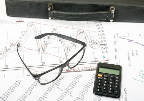 Glasses and suitcase with calculator on grpahical charts background, business concept