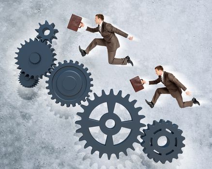 Two businessmen running on wheel gears on grey wall background