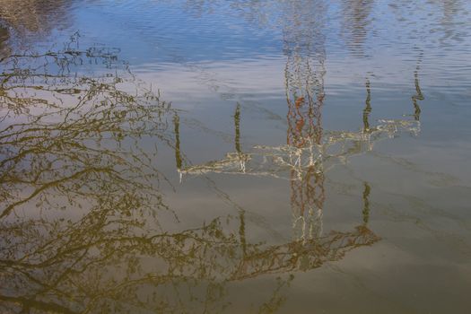 Surface of the water in the lake with small waves, reflecting the electricity tower, sky and twigs of a tree.