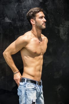 Handsome unshaven shirtless young man standing against black wall, looking to a side