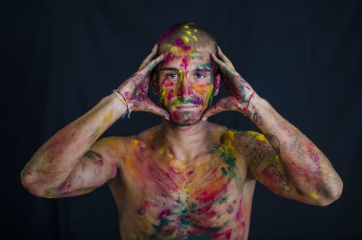 Head and shoulders shot of attractive young man shirtless, skin painted all over with bright Holi colors, looking at camera, isolated on white background