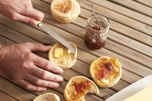 Hands spreading butter and strawberry marmalade on toasted english muffins with a knife