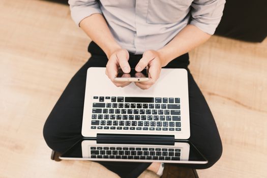 Top view man typing text message on his smart phone while sitting front open laptop computer, office man connecting to wireless via notebook and mobile phone while he keyboarding .