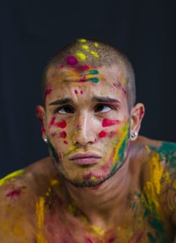 Head and shoulders shot of attractive young man shirtless, skin painted all over with bright Holi colors doing silly face expression, isolated on black background