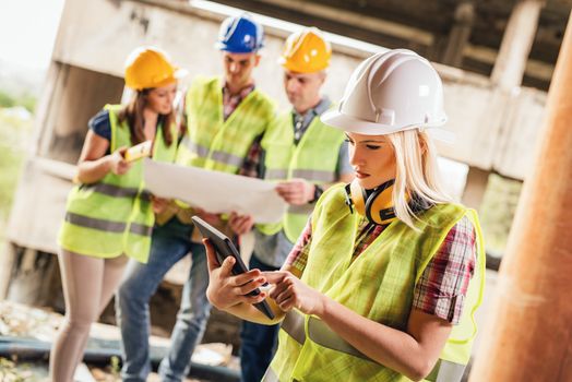 Beautiful young female construction architects using digital tablet at a construction site. Her colleagues review plan in background.