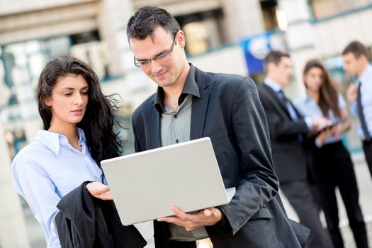 Smiling young businessman and businesswoman in front of office building looking at the laptop that holds a businessman.