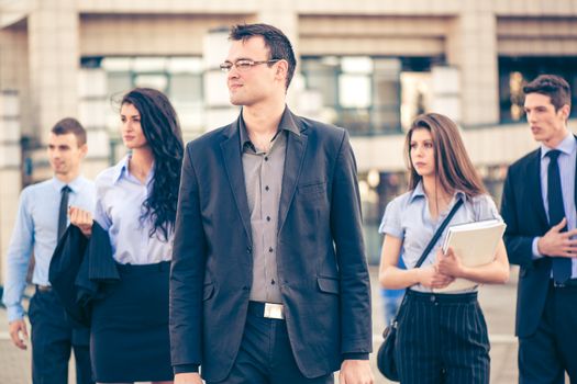 Young businessman, elegantly dressed, standing  with his team in front of office building. Everyone is looking to the side.