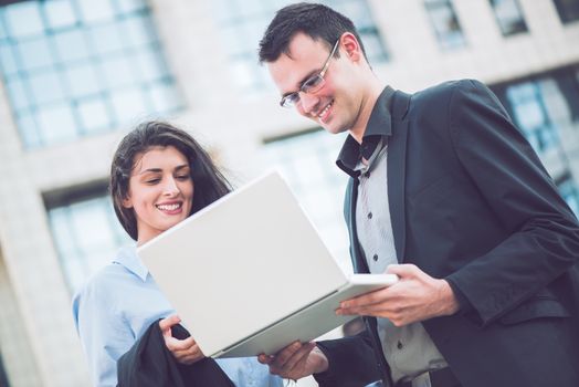 Smiling young businessman and businesswoman in front of office building looking at the laptop that holds a businessman.