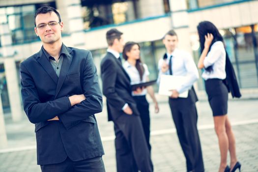 Young businessman standing in front of office building separated from the rest of the business team. With a smile looking at the camera.