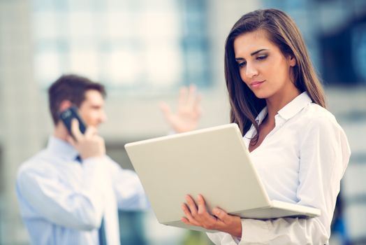 Young business partners standing in front of the building. Business woman working on a laptop while her partner use a mobile phone for business conversation.