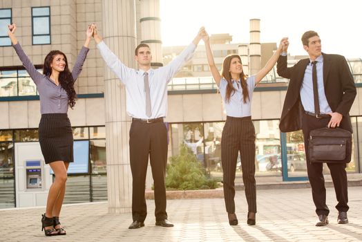 Young successful business people are hold with arms raised in front of the office building while the sun shines from behind.