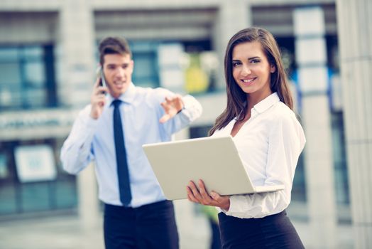 Young business partners standing in front of the building. Business woman working on a laptop while her partner use a mobile phone for business conversation.