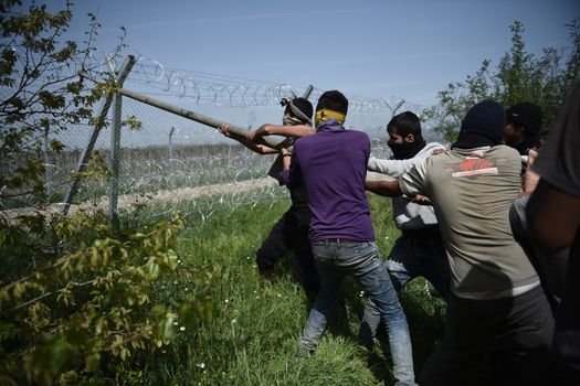 GREECE, Idomeni: Migrants and refugees try to break down the border fence near their makeshift camp at the Greek-Macedonian border near Idomeni village, on April 13, 2016 as Macedonian police fire tear gas cans. About 100 migrants spread out over about 100 metres (yards) tugging at the wire fence, but swiftly pulled back when two squads of Greek riot police moved in, the reporter said. The Greek riot police positioned themselves between the migrants and the Macedonian fence, ending the incident.