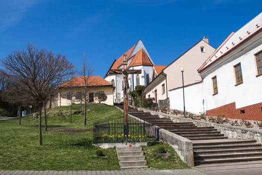 Traditional slovak village, built on a hill, with a church on the top of it. Wooden cross beside the stairs to the gothic church. Springtime with a green grass and blue sky.