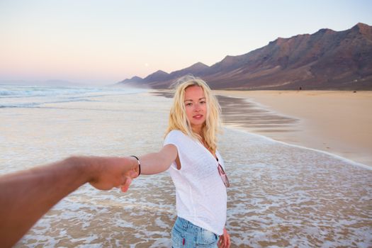 Young romantic couple, holding hands, having fun on perfect deserted beach at sunset. Shot from boyfrieds perspective. Guy looking at her beautiful carefree girlfriend.