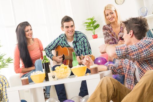 A small group of young people hang out at the house party, chatting with each other while their friend having fun playing acoustic guitar.