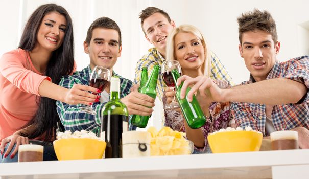 Cheerful friends at home party toasting knocking with bottles and glasses. With a smile looking at the camera.