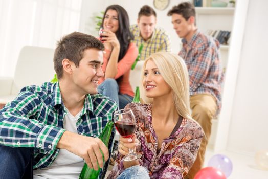 Young couple at a home party, sit on the floor, knocking the glasses toasting, in the background you can see friends.