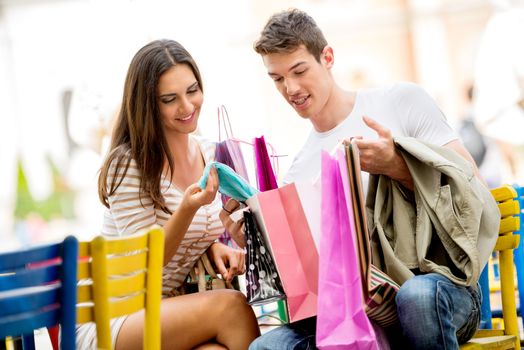 Girl and her boyfriend are resting after shopping in outdoor cafe with colorful chairs, watching with a smile in shopping bags, excited about the things they bought.