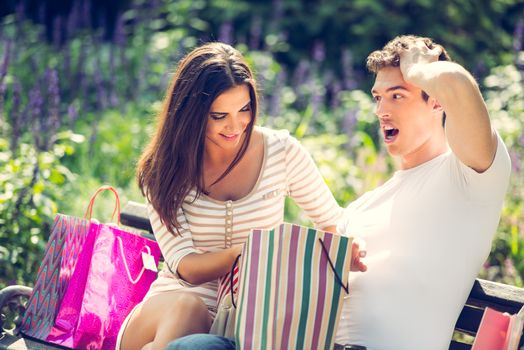 Young guy with a worried expression on his face and his smiling girlfriend who sits on a park bench next to a bunch of shopping bags.