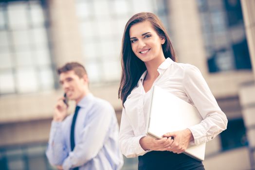 Young business partners standing in front of the building. Business woman carrying laptop while her partner use a mobile phone for business conversation.