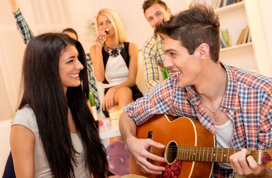 A young man with an acoustic guitar, at home party, courting a pretty girl playing to her. In the background you can see young people sitting on the couch and enjoy the atmosphere of home party.