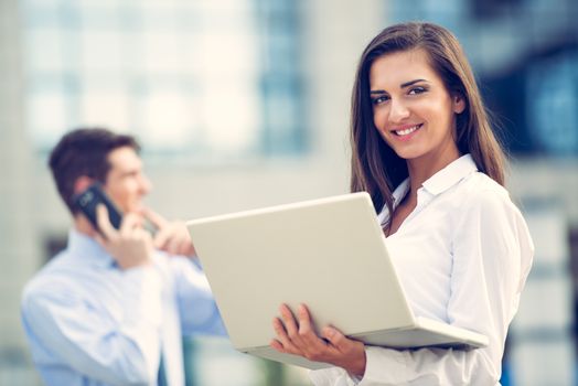 Young business partners standing in front of the building. Business woman working on a laptop while her partner use a mobile phone for business conversation.